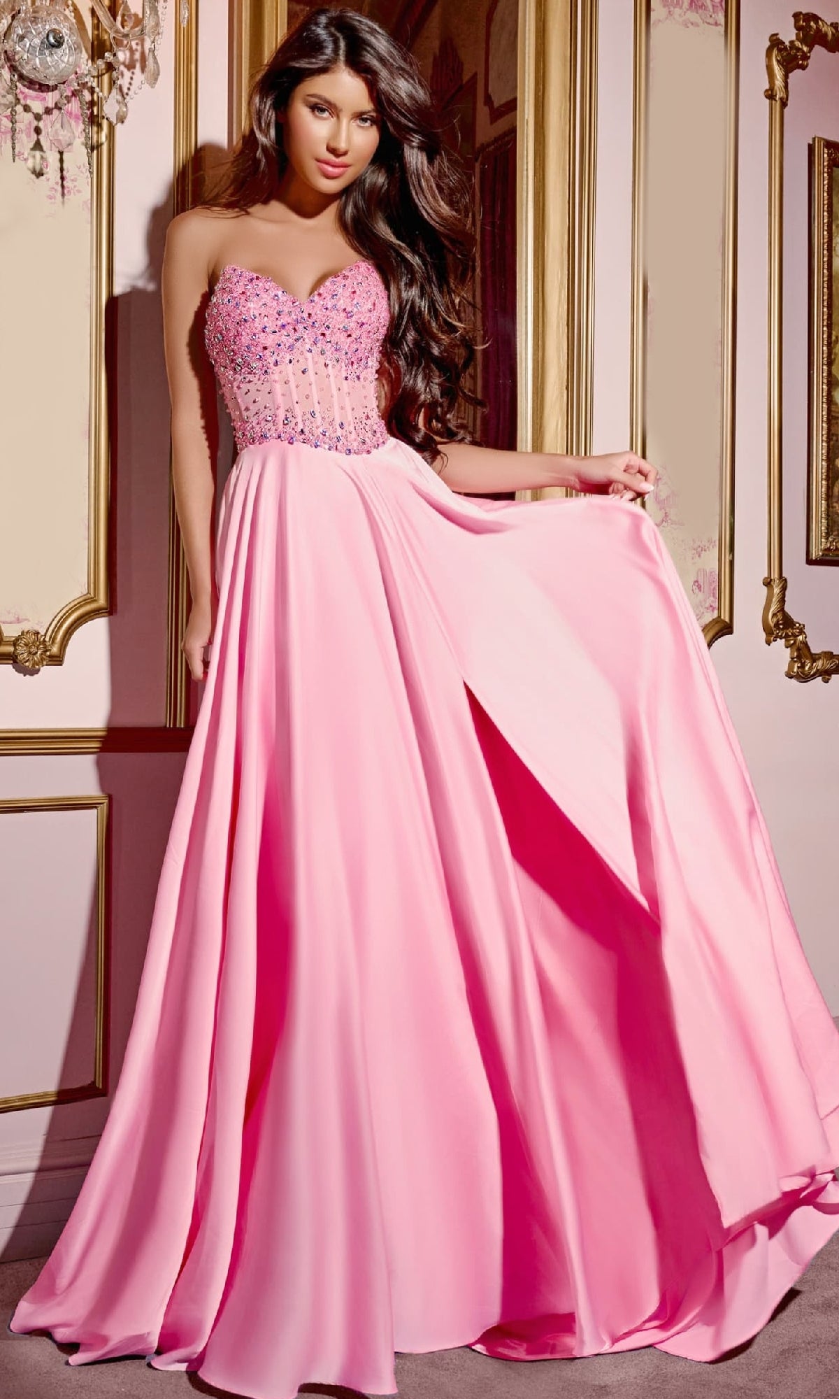 A woman wearing the Jovani D6021 Pink Long Prom Dress with a beaded corset bodice and flowing A-line skirt stands indoors against ornate, gold-trimmed walls.